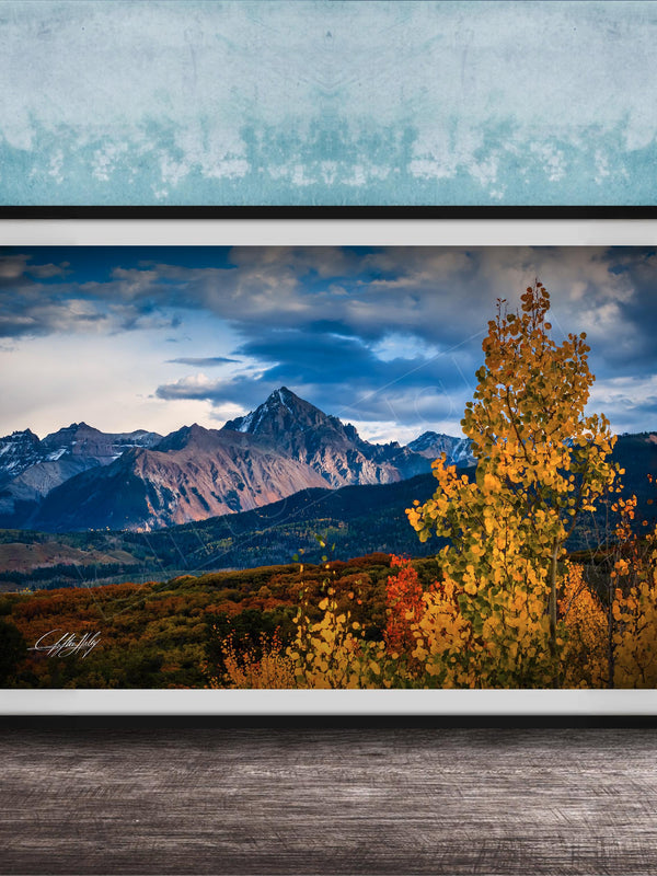 The image is a framed photograph depicting a scenic mountain landscape with a mix of autumnal and winter colors, including vibrant yellow and orange foliage in the foreground and majestic snow-capped peaks in the background.