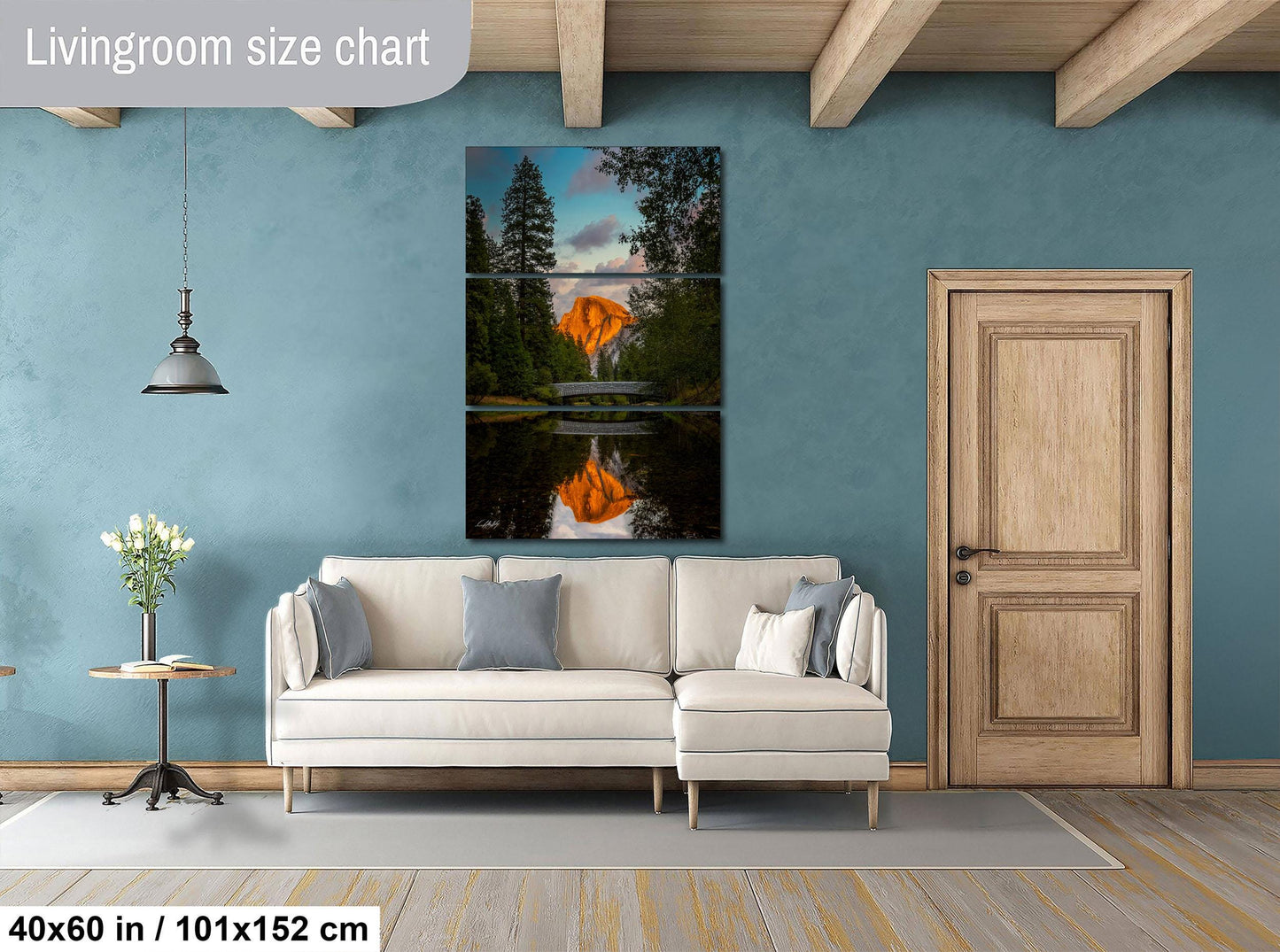 The image is a living room with a large painting of a sunset over a lake hanging on the wall. The painting is divided into three panels, with the top and bottom panels depicting the sunset and the middle panel showing the reflection of the sunset on the water. The room has a white sofa, a wooden door, and a small table with a vase of flowers. The wall is painted in a light blue color, and the floor is made of wood.