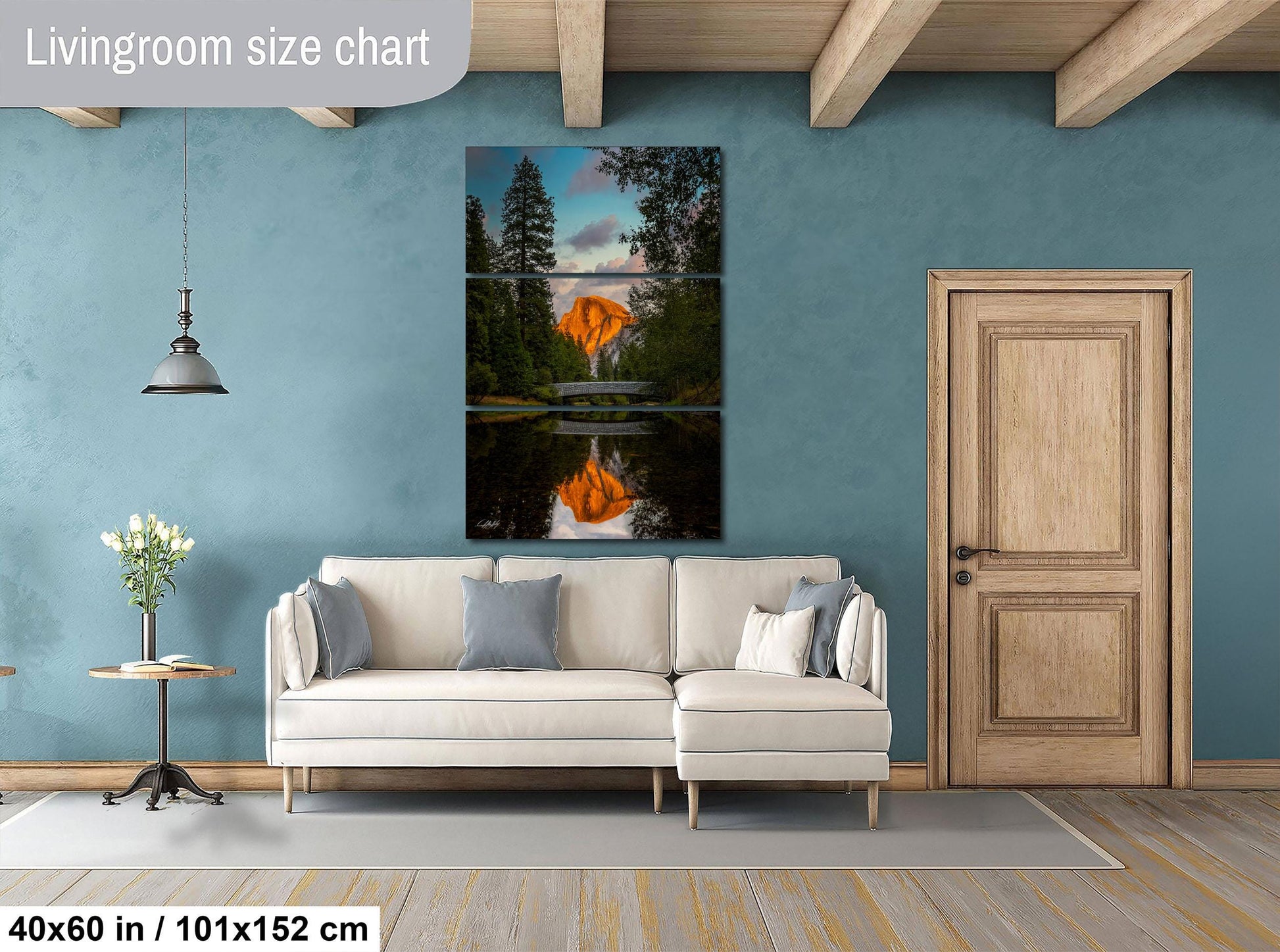 The image is a living room with a large painting of a sunset over a lake hanging on the wall. The painting is divided into three panels, with the top and bottom panels depicting the sunset and the middle panel showing the reflection of the sunset on the water. The room has a white sofa, a wooden door, and a small table with a vase of flowers. The wall is painted in a light blue color, and the floor is made of wood.