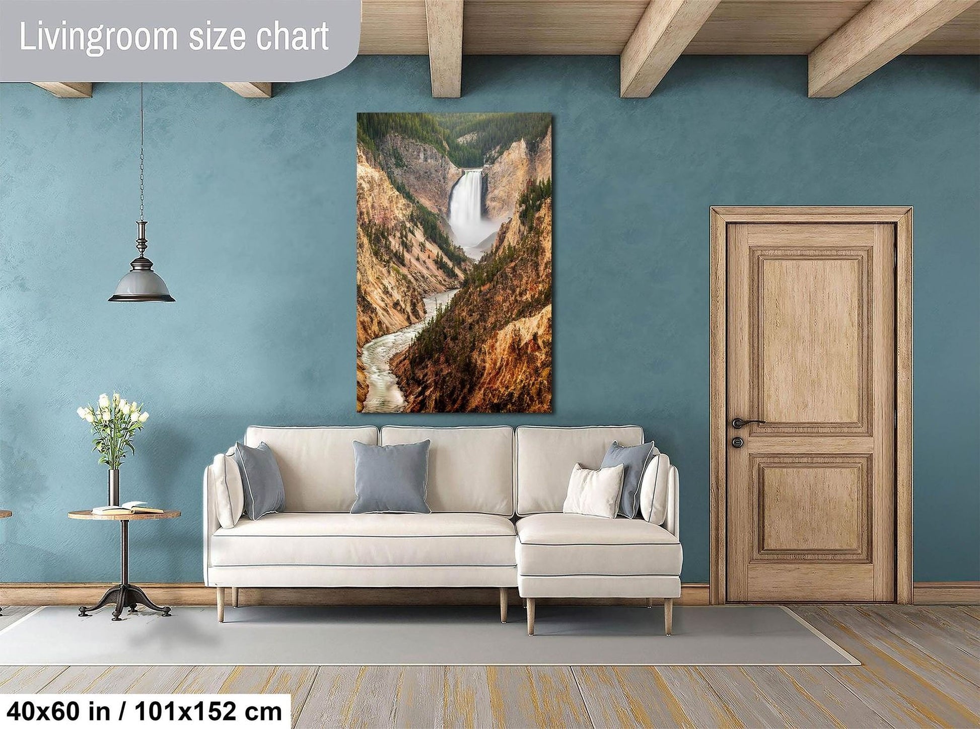 a living room with a large painting of a waterfall hanging on the wall. The painting is framed in a white frame and is approximately 40 inches wide and 51 inches tall. The room has a white sofa, a wooden door, and a wooden table with a vase of flowers. The walls are painted a light blue color, and the floor is made of wood.