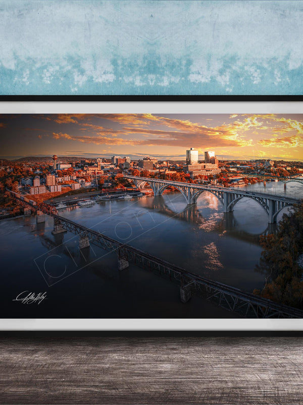 The image is a framed photograph depicting a cityscape with a bridge spanning a river, set against a backdrop of a colorful sunset sky.