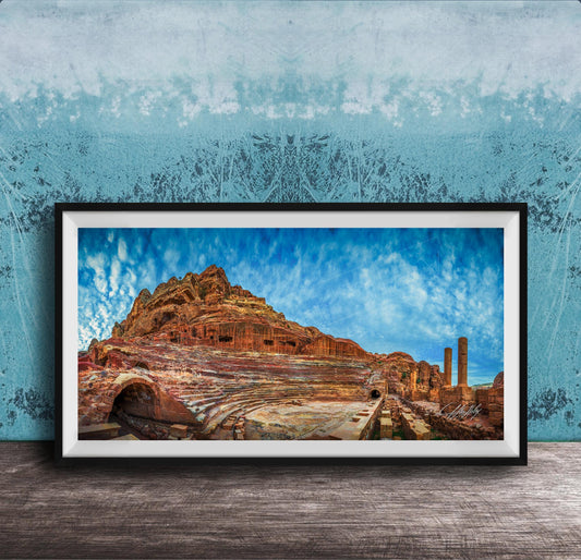 The image is a framed photograph of a desert landscape with a rocky hillside, a cave, and a few buildings or structures in the background.