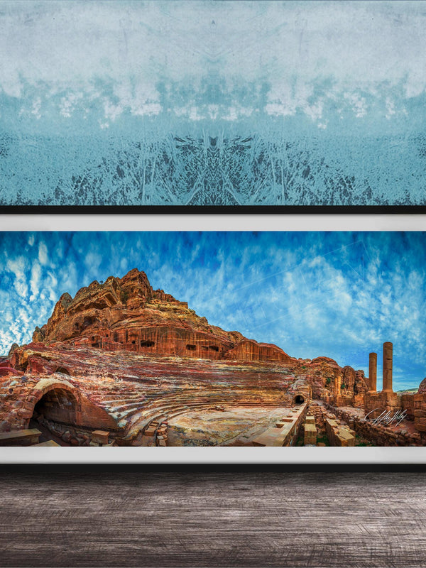The image is a framed photograph of a desert landscape with a rocky hillside, a cave, and a few buildings or structures in the background.