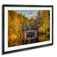 A framed photograph depicting a stone bridge surrounded by a forest with autumn-colored trees reflected in the water below.