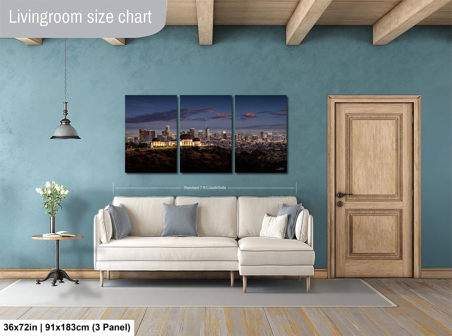 The image is a living room with a large wall-mounted painting depicting a city skyline at night. The painting is divided into three panels, with the largest panel covering the entire wall. The room has a white sofa, a wooden door, and a small table with a vase of flowers. The painting is approximately 36 inches wide and 15 inches tall, and it is mounted on a blue wall.