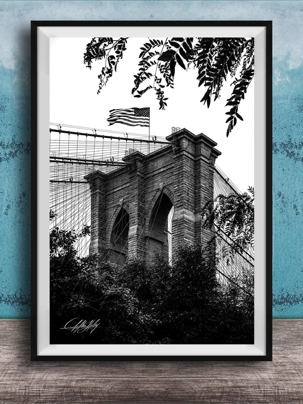 A black and white photograph of the Brooklyn Bridge, with the American flag flying atop the structure.