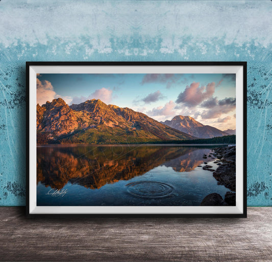 A framed photograph depicting a serene mountain landscape with a calm lake reflecting the vibrant colors of the sky and surrounding mountains.