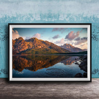 A framed photograph depicting a serene mountain landscape with a calm lake reflecting the vibrant colors of the sky and surrounding mountains.