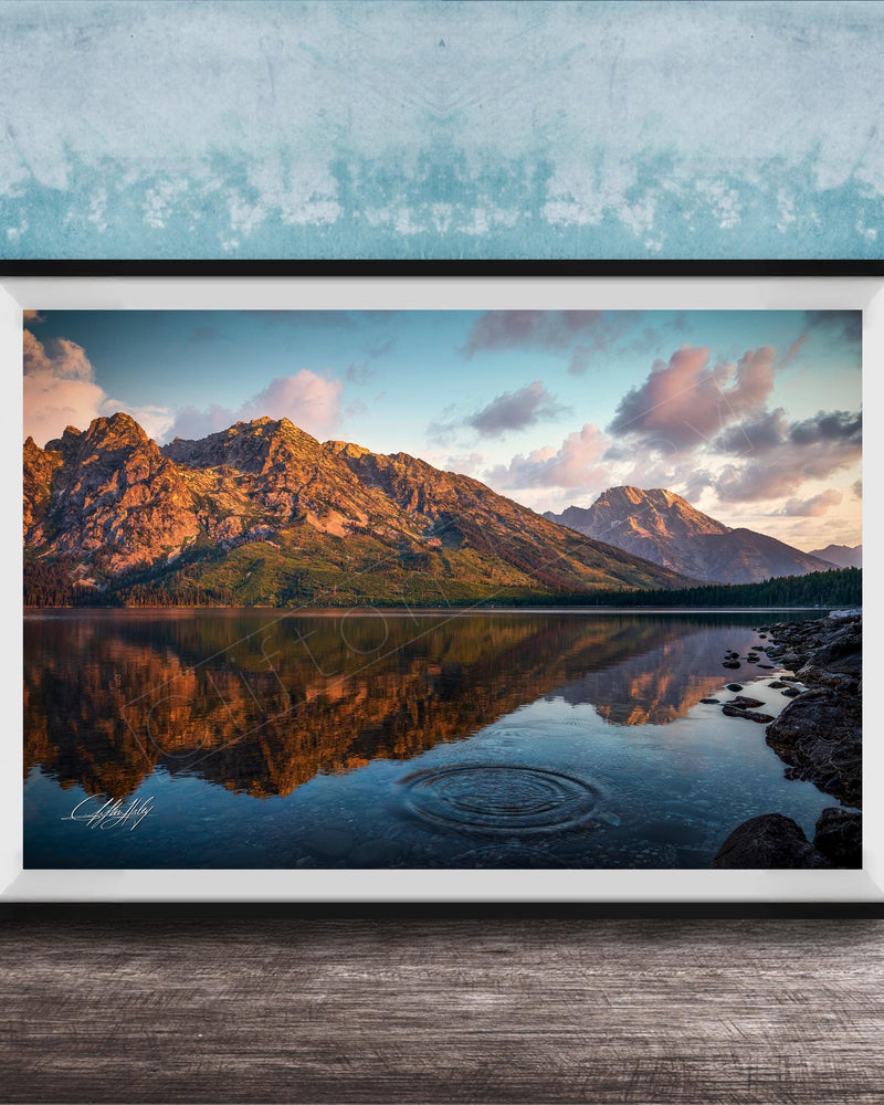 A framed photograph depicting a serene mountain landscape with a calm lake reflecting the vibrant colors of the sky and surrounding mountains.