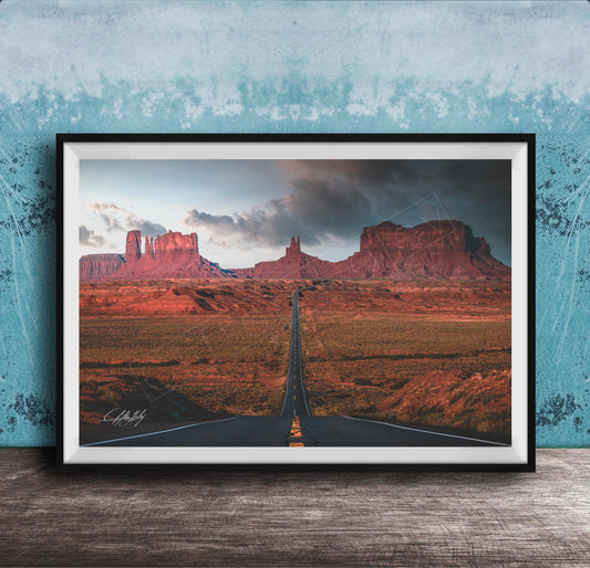 The image is a framed photograph depicting a desert landscape with a winding road leading towards a series of red rock formations in the distance.
