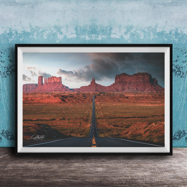 The image is a framed photograph depicting a desert landscape with a winding road leading towards a series of red rock formations in the distance.