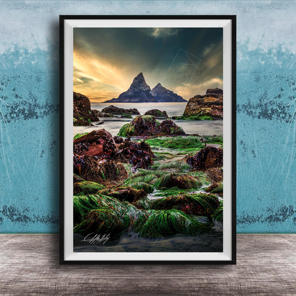The image is a framed photograph of a rocky shoreline with a large, distant mountain in the background. The photograph is set against a blue wall, and the sky above is cloudy. The image is a collage of multiple images, and the photograph is framed with a black border.