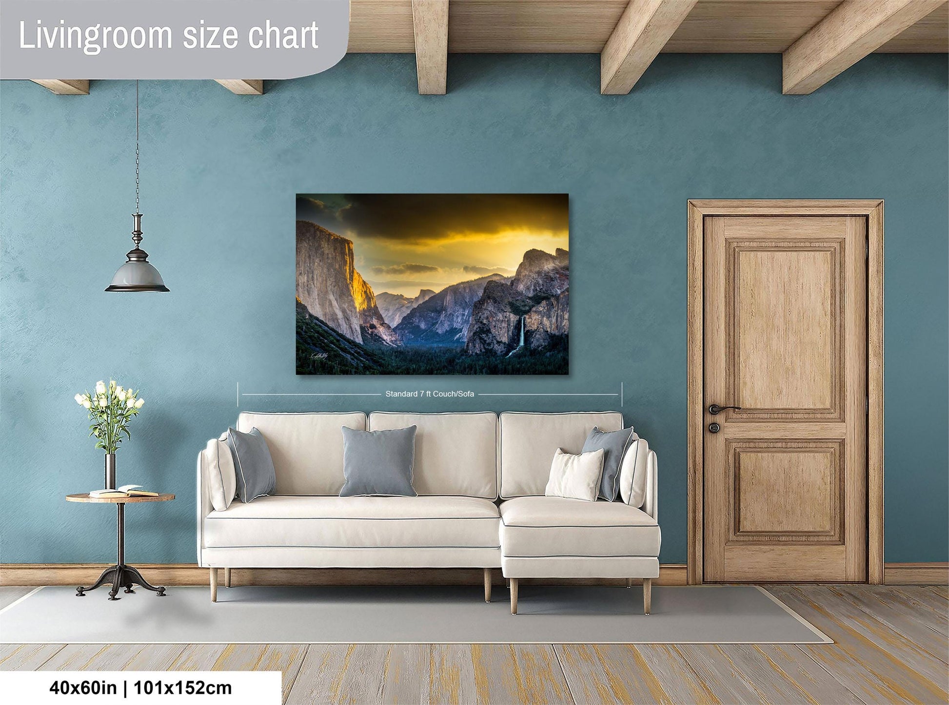 a living room with a large wall painting of a mountainous landscape, a white sofa, a wooden door, and a small table with a vase of flowers.