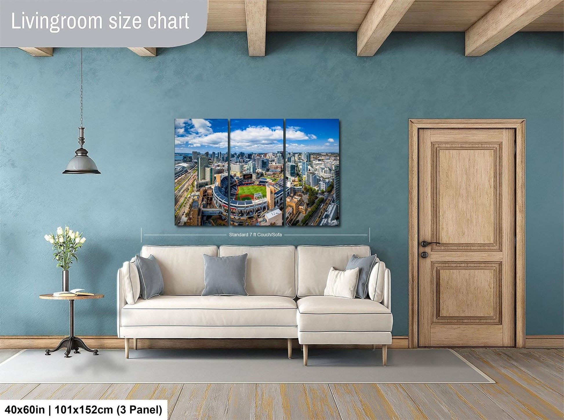 The image is a living room with a large, high-resolution painting of a cityscape hanging on the wall. The painting is divided into three panels, depicting a panoramic view of the city. The room has a white sofa, a wooden door, and a small table with a vase of flowers. The painting is approximately 40 inches wide and 101.5 inches tall, with a 3-panel layout.