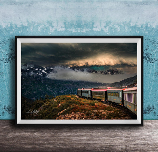 A framed photograph depicting a train traveling through a mountainous landscape with dramatic clouds and a blue sky in the background.