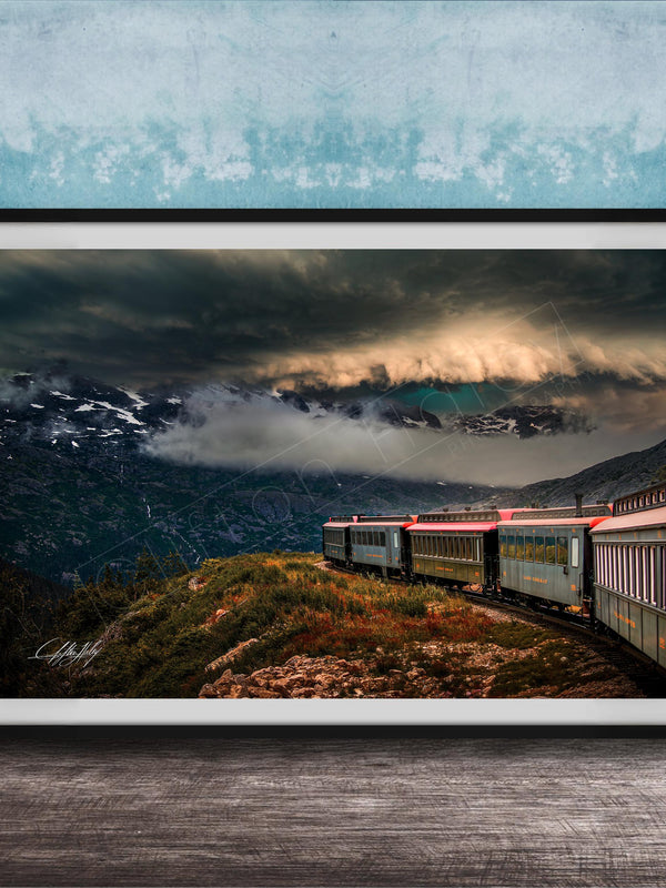 A framed photograph depicting a train traveling through a mountainous landscape with dramatic clouds and a blue sky in the background.