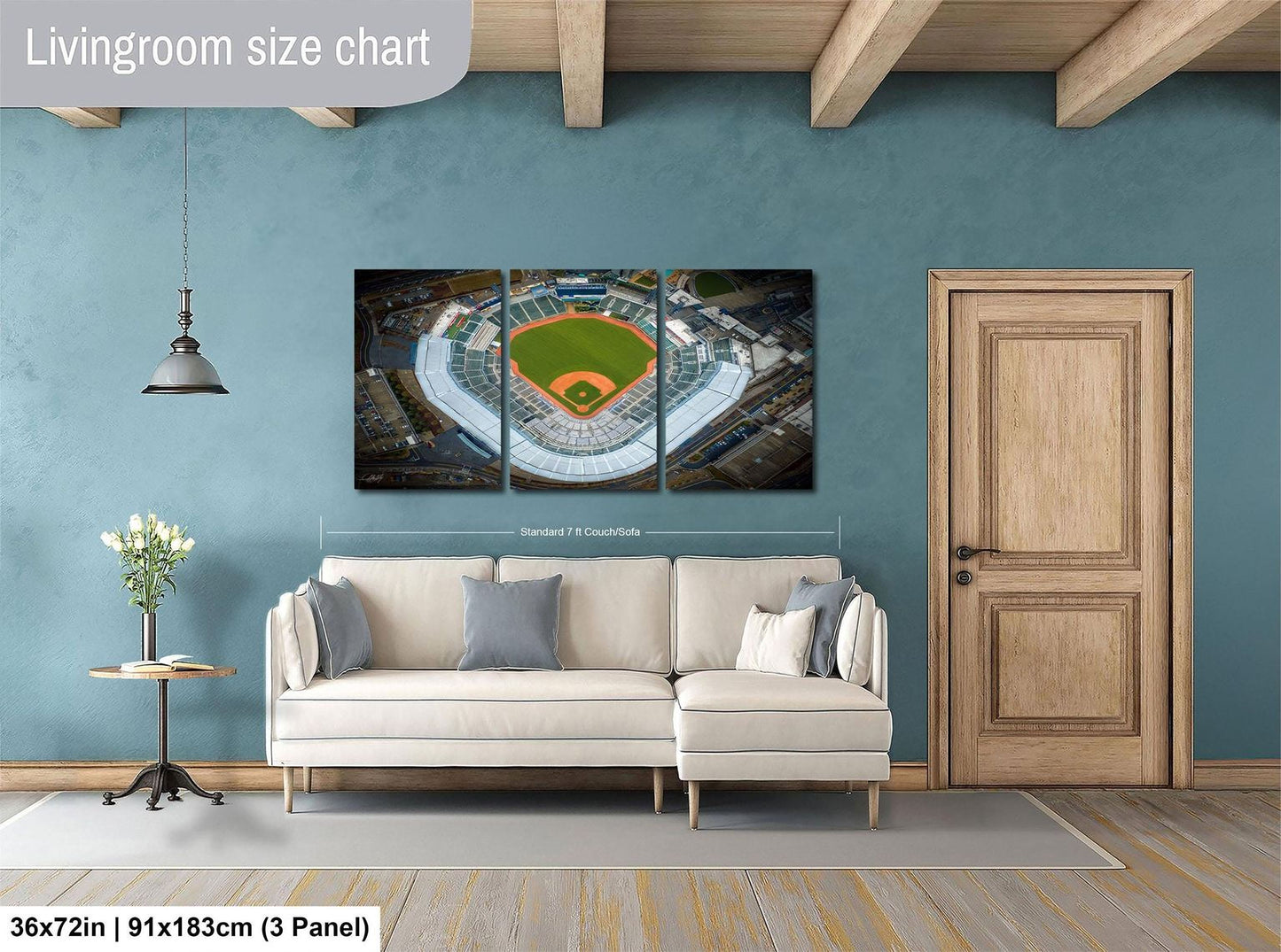 The image is a living room with a large wall art depicting an aerial view of a baseball stadium. The wall art is divided into three panels, with the largest panel covering the entire wall. The room is furnished with a white sofa and a wooden door, and there is a potted plant and a vase on a table. The wall art is approximately 36 inches wide and 9 inches tall, and it is mounted on a wooden beam.
