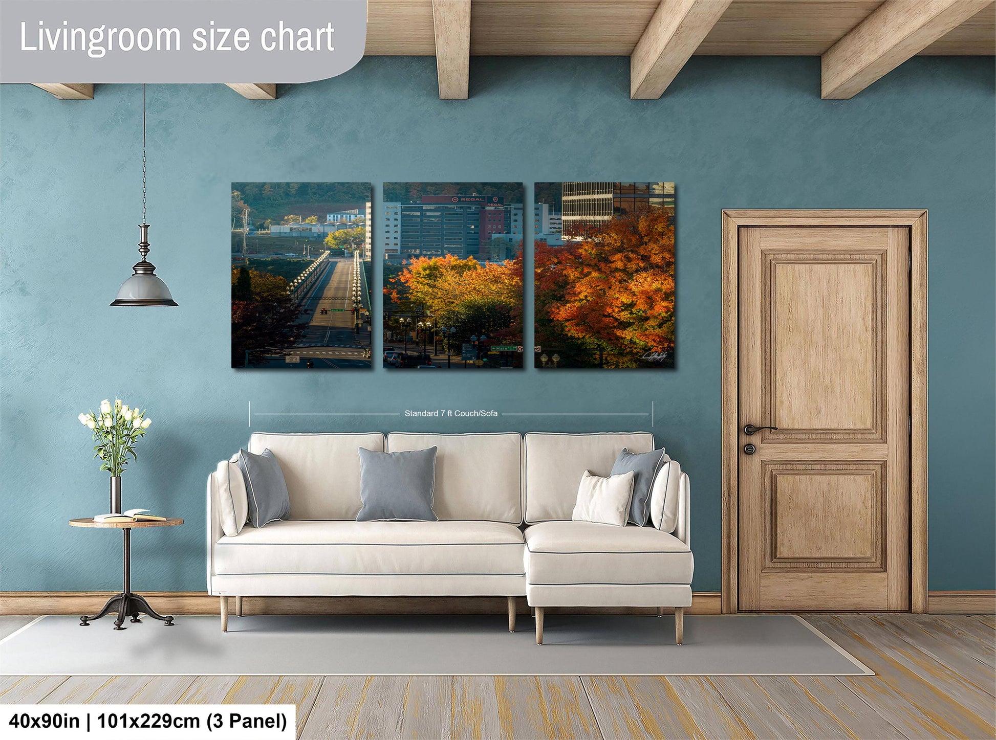 The image is a living room with a white sofa and a wooden door. Above the sofa, there is a large wall art panel depicting a cityscape with a bridge and trees. The wall art is framed by a wooden border and is approximately 4 feet wide.