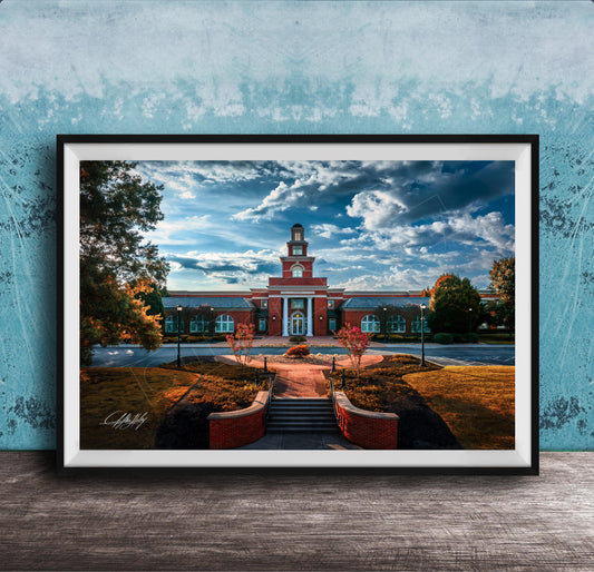 A framed photograph of a red brick building with a clock tower, surrounded by trees and a brick pathway leading up to it, set against a blue sky with clouds.