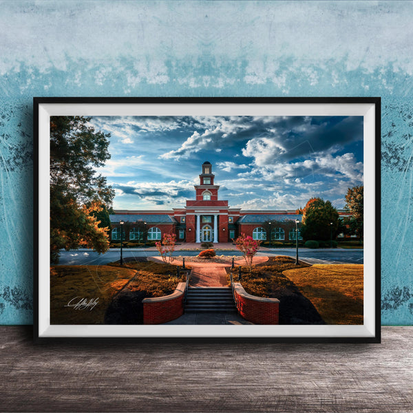 A framed photograph of a red brick building with a clock tower, surrounded by trees and a brick pathway leading up to it, set against a blue sky with clouds.