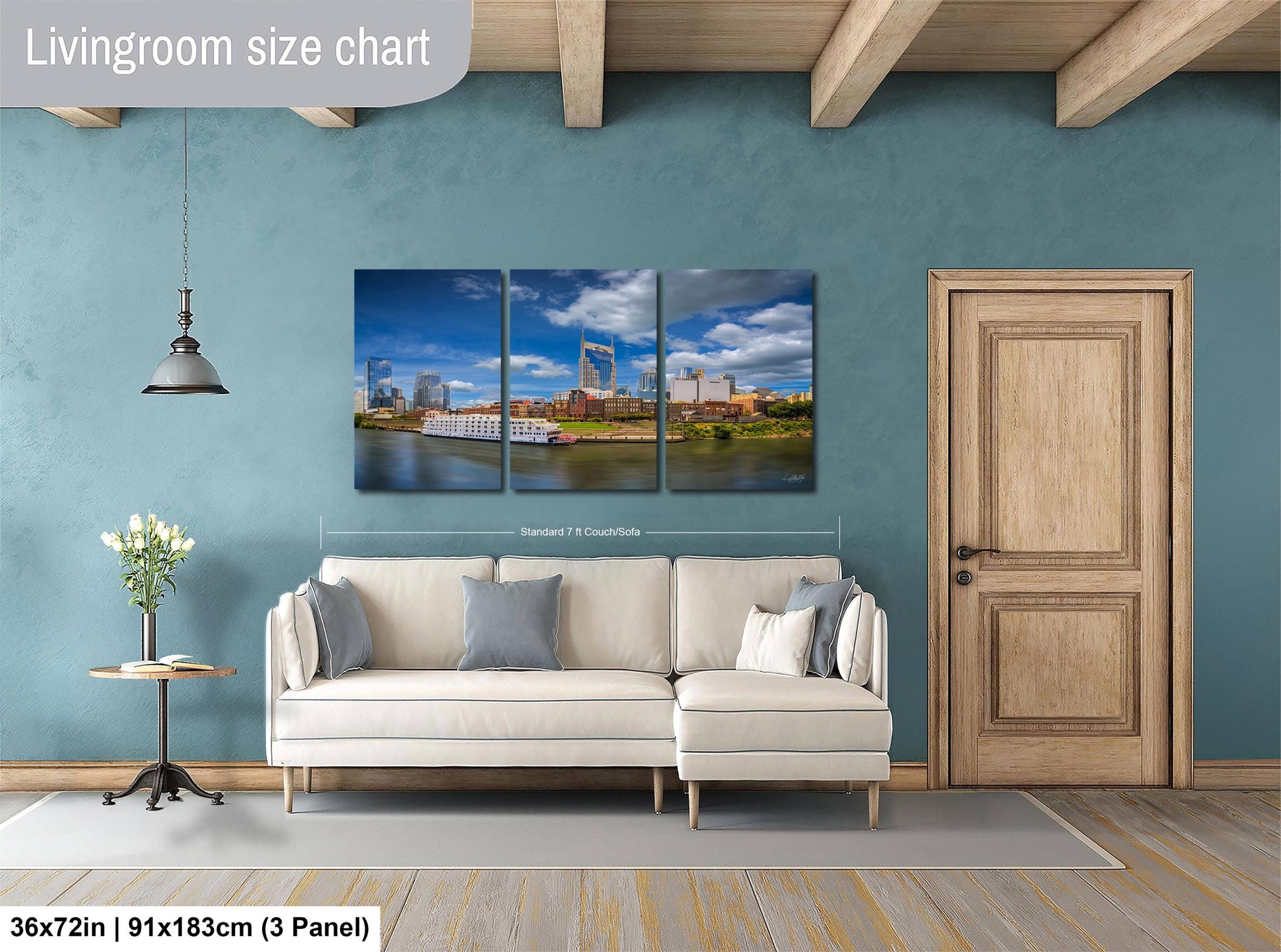 The image is a living room with a large wall art depicting a cityscape. The wall art is divided into three panels, each showing a different part of the city. The living room has a white sofa and a wooden door, and there is a potted plant and a vase on a table nearby. The wall art is approximately 36 inches wide and 9 inches tall, and it is mounted on a blue wall.
