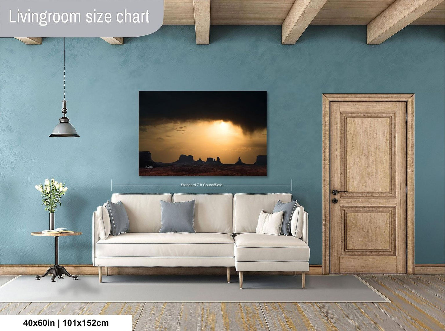 a living room with a large wall painting depicting a desert landscape at sunset. The painting is framed and hangs above a white sofa with two gray pillows. The room has a wooden door and a small table with a vase of flowers. The wall is painted in a light blue color, and the floor is covered with a gray rug.
