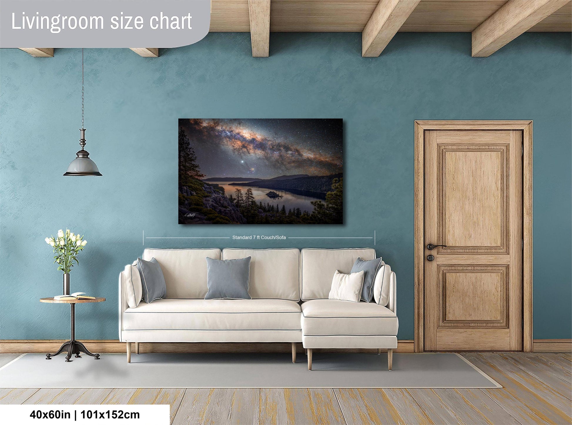a living room with a large wall painting of a serene landscape featuring a lake and mountains. The painting is hung above a white sofa, which is positioned on a gray rug. The room has a wooden door and a small table with a vase of flowers. The wall painting is approximately 40 inches wide and 115 centimeters tall.