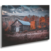 A rustic wooden barn stands in a field, surrounded by trees with vibrant orange leaves against a cloudy sky.