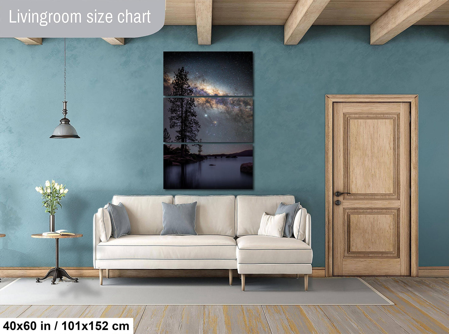 The image is a living room with a large, three-piece wall art depicting a night sky with a galaxy. The wall art is mounted above a white sofa, and there is a wooden door and a small table with a vase of flowers nearby. The room has a blue wall and wooden beams, creating a cozy and inviting atmosphere.