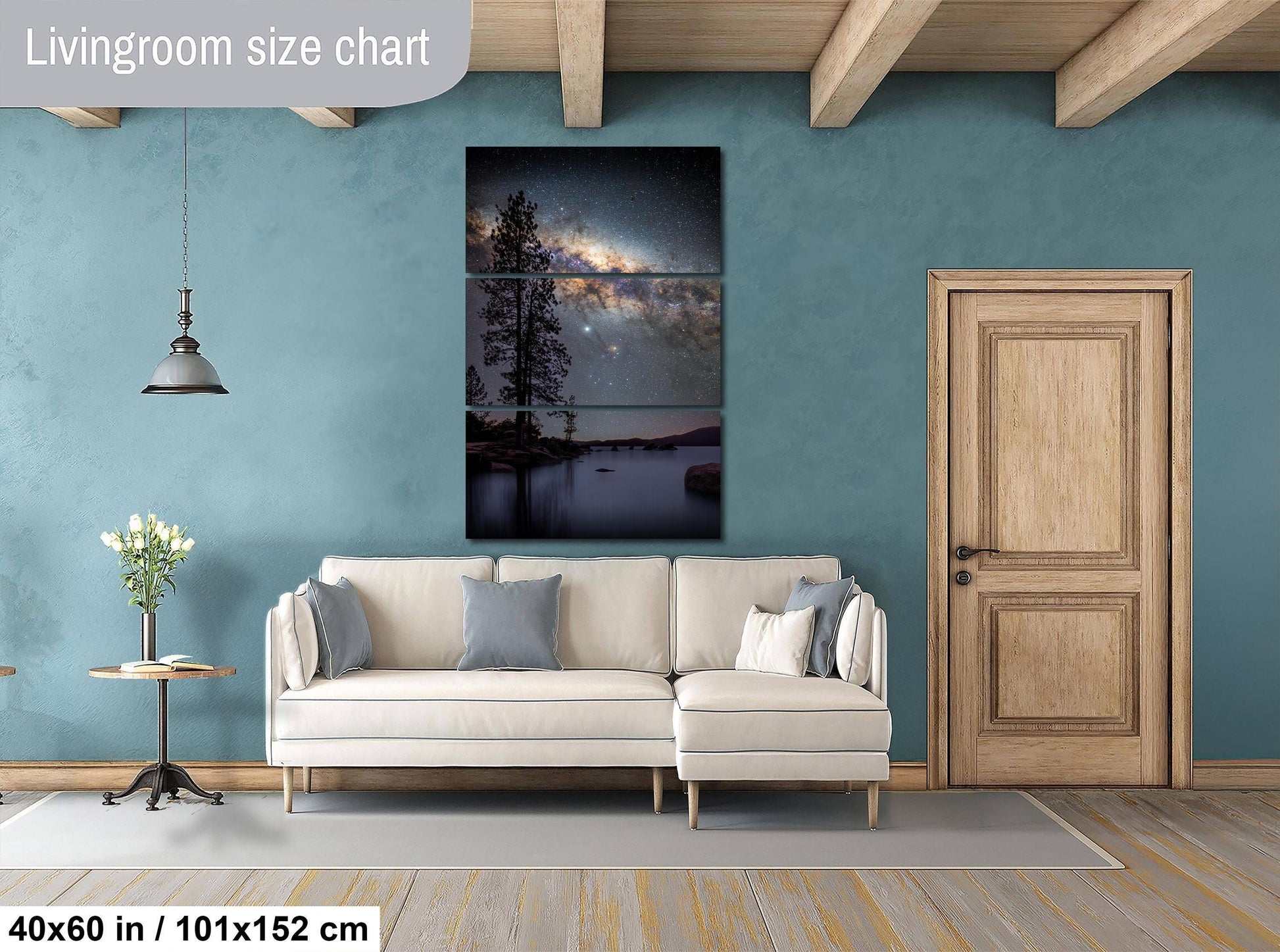 The image is a living room with a large, three-piece wall art depicting a night sky with a galaxy. The wall art is mounted above a white sofa, and there is a wooden door and a small table with a vase of flowers nearby. The room has a blue wall and wooden beams, creating a cozy and inviting atmosphere.