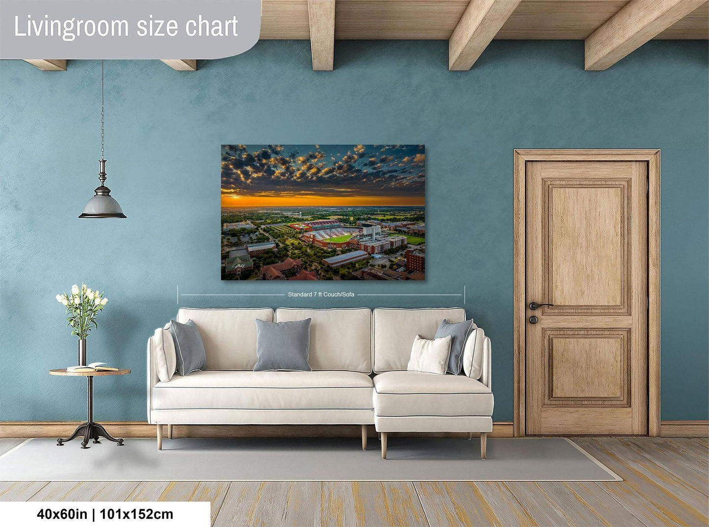 a living room with a large wall-mounted painting depicting a cityscape at sunset. The painting is framed and hangs above a white sofa. The room has a wooden door and a small table with a vase of flowers. The wall has a blue color, and the floor is made of wood.