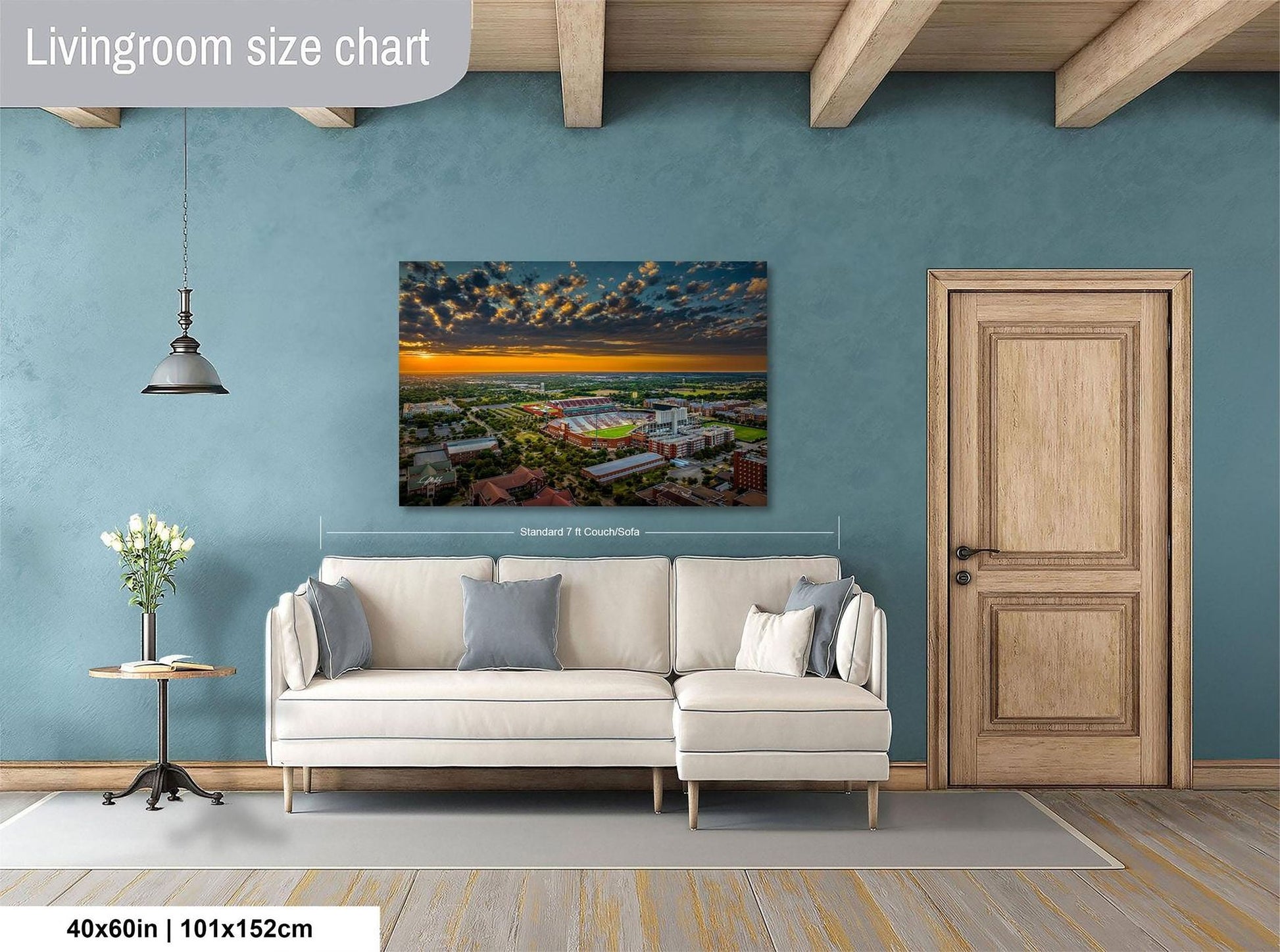 a living room with a large wall-mounted painting depicting a cityscape at sunset. The painting is framed and hangs above a white sofa. The room has a wooden door and a small table with a vase of flowers. The wall has a blue color, and the floor is made of wood.