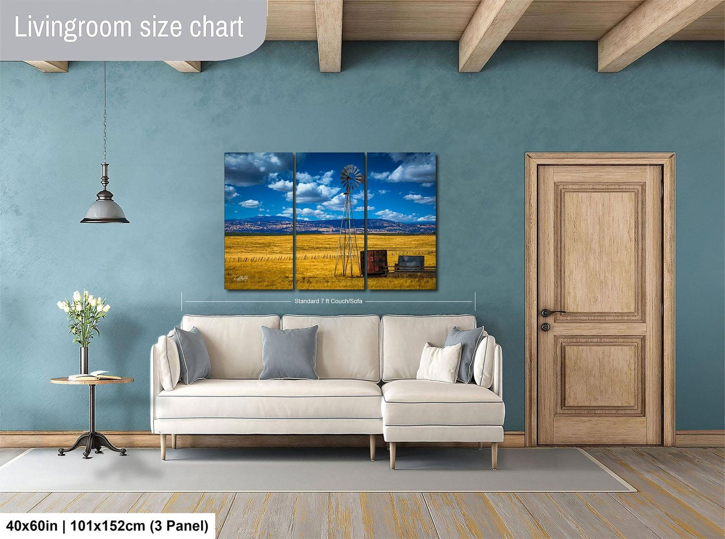 The image is a living room with a large painting of a windmill and a field in the background. The painting is mounted on the wall above a white sofa. The room has a wooden door and a wooden beam ceiling.