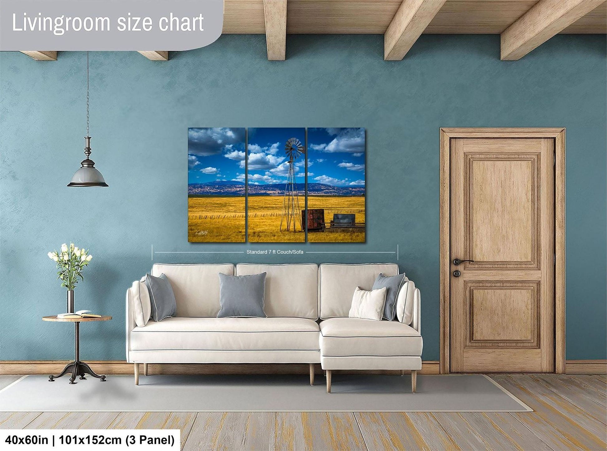 The image is a living room with a large painting of a windmill and a field in the background. The painting is mounted on the wall above a white sofa. The room has a wooden door and a wooden beam ceiling.