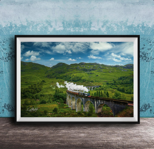 A framed photograph depicting a scenic train traveling over a stone bridge in a lush, green landscape with rolling hills and a blue sky with clouds.