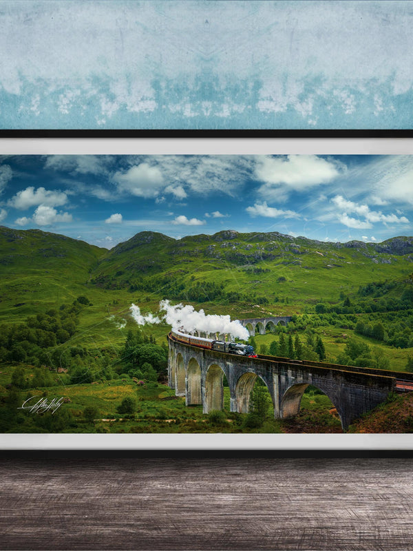 A framed photograph depicting a scenic train traveling over a stone bridge in a lush, green landscape with rolling hills and a blue sky with clouds.
