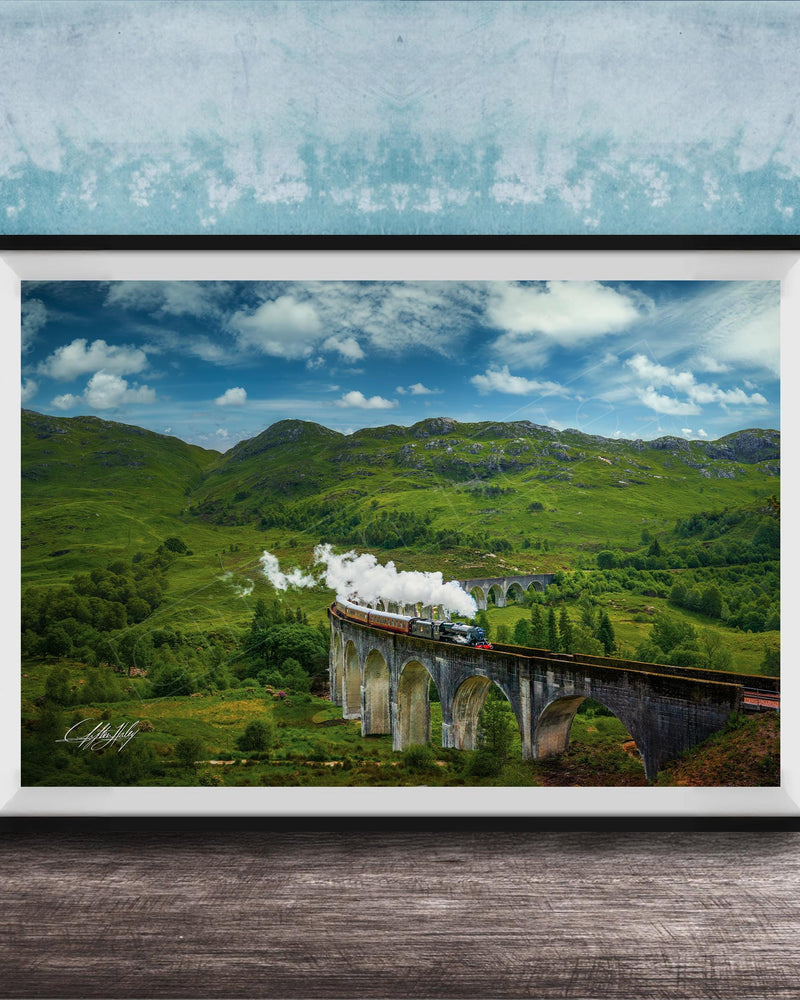A framed photograph depicting a scenic train traveling over a stone bridge in a lush, green landscape with rolling hills and a blue sky with clouds.