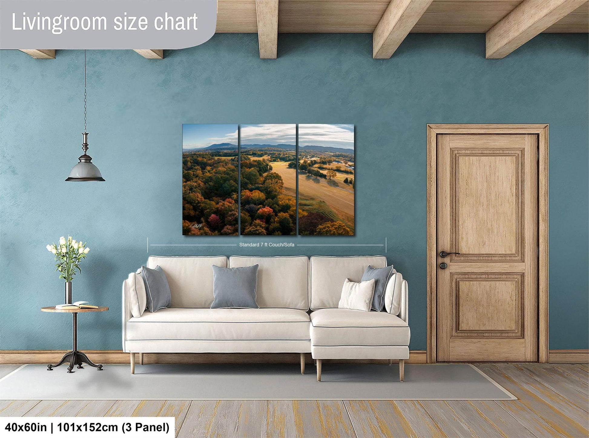 The image is a living room with a large, three-panel landscape painting hanging on the wall. The painting depicts a scenic countryside view with rolling hills, trees, and a winding road. The room is furnished with a white sofa and a wooden door, creating a cozy and inviting atmosphere.