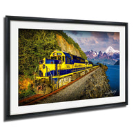 A yellow and blue train engine, numbered 3001, is traveling along a track that runs along the edge of a cliff overlooking a body of water and snow-capped mountains in the distance.