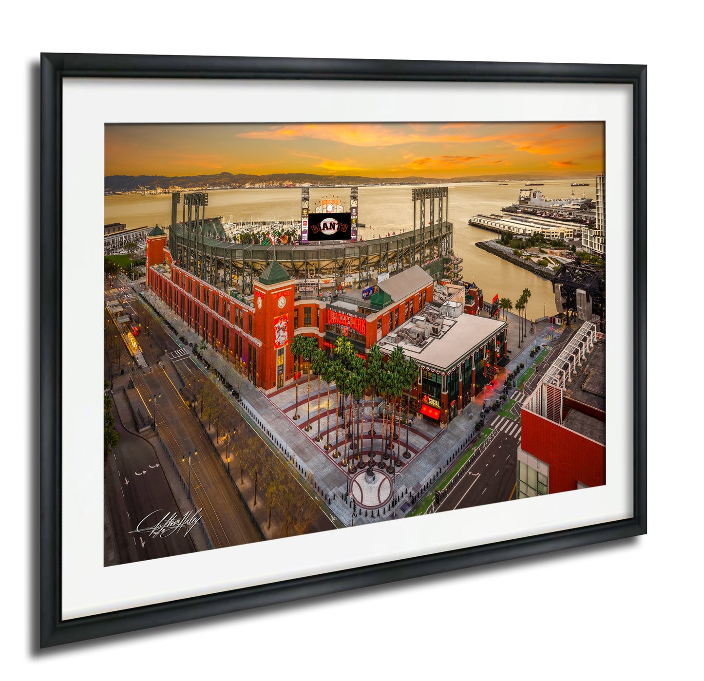 The image is a framed aerial view of a large stadium with a red and white exterior, surrounded by palm trees and other buildings. The stadium appears to be a sports venue, with a large screen visible in the background. The sky is filled with warm, orange and yellow hues, suggesting a sunset or sunrise. The image is framed with a black border, and the artist's signature is visible in the bottom left corner.