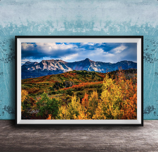 The image is a framed photograph of a mountainous landscape with autumn-colored trees in the foreground and a blue sky with clouds in the background.