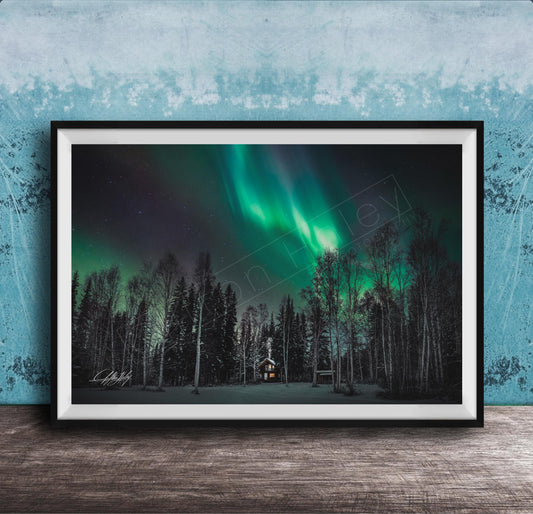 A framed photograph depicting a serene night scene with a forest and a house illuminated by the vibrant green and blue hues of the Aurora Borealis ( Aurora lights) in the sky.