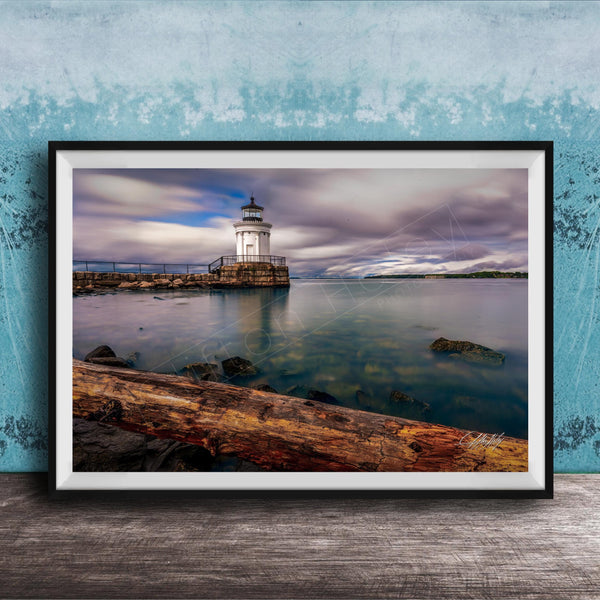 A framed photograph of a lighthouse on a rocky shore, with a calm body of water in the background and a cloudy sky above.