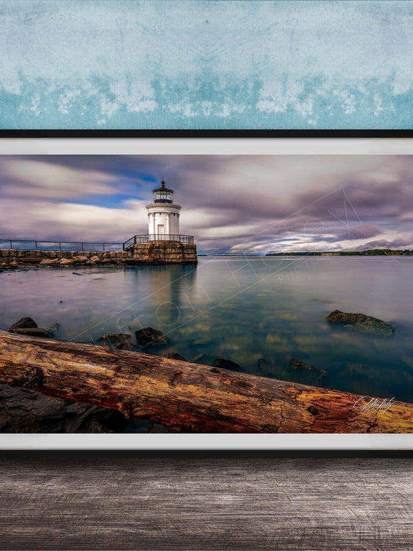 A framed photograph of a lighthouse on a rocky shore, with a calm body of water in the background and a cloudy sky above.