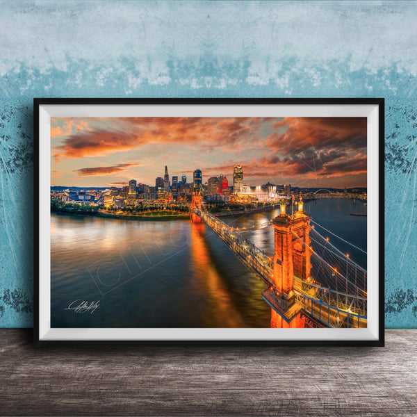 A framed photograph of the Cincinnati skyline at sunset, with the city's iconic landmarks and bridges illuminated against a dramatic sky.