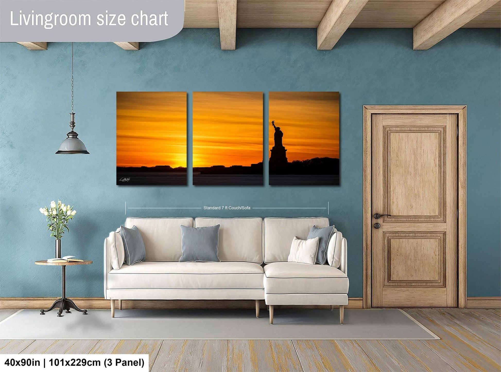 The image is a living room with a white sofa and a wooden door. Above the sofa, there is a large wall art piece depicting a sunset over a silhouette of a person. The wall art is composed of three panels, each showing a different stage of the sunset. The living room has a blue wall and a wooden floor, creating a cozy and inviting atmosphere.