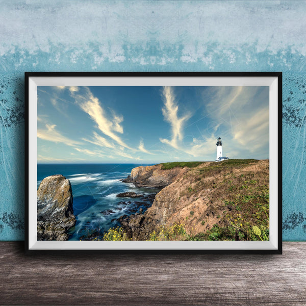 A framed photograph of a lighthouse on a cliff overlooking the ocean, with a cloudy blue sky and crashing waves in the foreground.