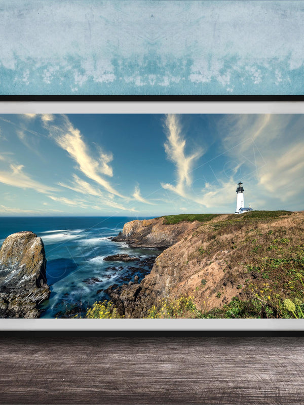 A framed photograph of a lighthouse on a cliff overlooking the ocean, with a cloudy blue sky and crashing waves in the foreground.