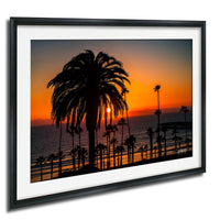 A framed photograph depicting a palm tree silhouetted against a vibrant sunset over the ocean.