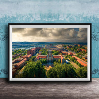 The image is a framed aerial view of a city with a mix of buildings, trees, and a river. The sky is cloudy, creating a dramatic backdrop for the cityscape.